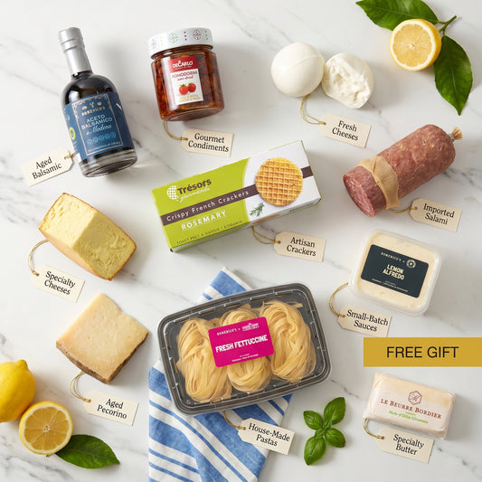 The DOM Society Summer Box collection on white marble with lemons and a blue striped linen: fresh buffalo mozzarella, smoked scamorza, aged pecorino, salami, rosemary crackers, fresh fettuccine, lemon alfredo sauce, semi-dried cherry tomatoes, aged balsamic of Modena, and Bordier butter, each with a kraft tag.